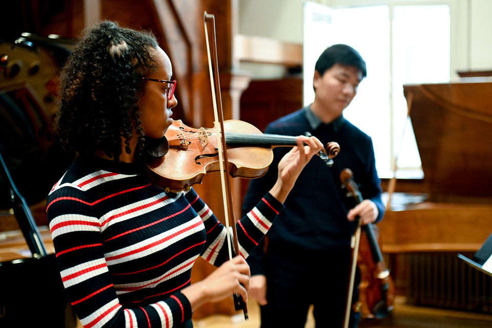 A student playing the violin, with a student holding a violin in the background